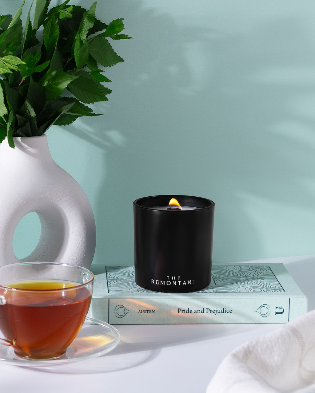 A black cylindrical candle with a lit flame sits on top of a light blue book. The candle has the words 'THE REMONTANT' printed on it. Remontant cozy kirkland candle