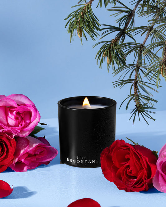 A black scented candle jar with a lit flame sits on a light blue surface, surrounded by red and pink roses and pine branches against a soft blue background. The candle label reads 'THE REMONTANT'.