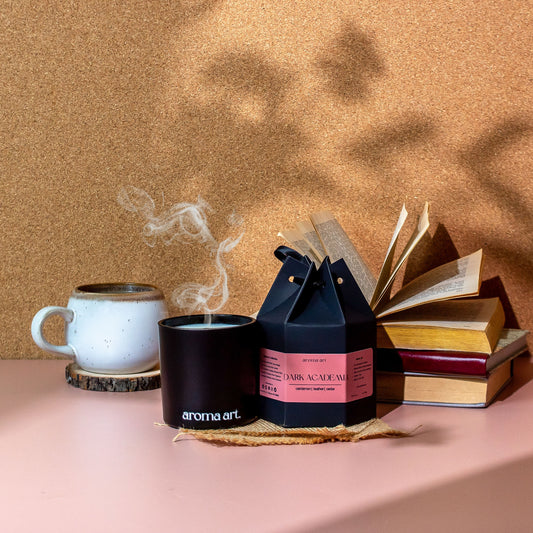 7oz crackling wooden wick soy coconut wax candle named Dark Academia, with notes of cardamon, leather, and cedar. A mysterious and elegant scent perfect for a night in with a good book. Shown with a cozy mug of tea and a stack of vintage books.