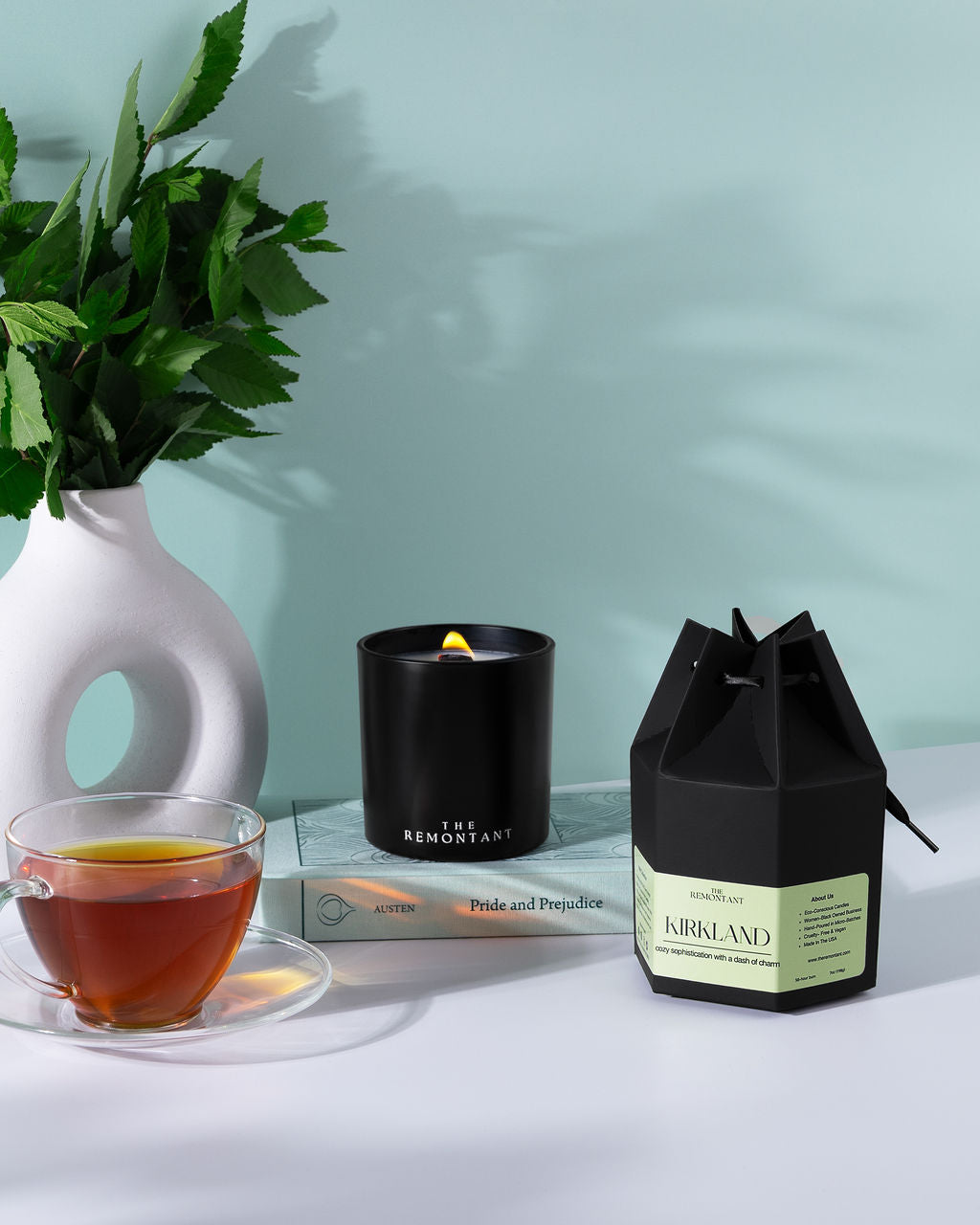 A styled product scene showing a black jar candle with a lit wick, a black gift box labeled kirkland candle  and a white vase with green leaves on a tabletop against a pale blue wall. A glass cup of tea is also present as a prop.This a Remontant " Gift Me " Candle in their iconic hexagonal gift box, classy, elegant , effortless.