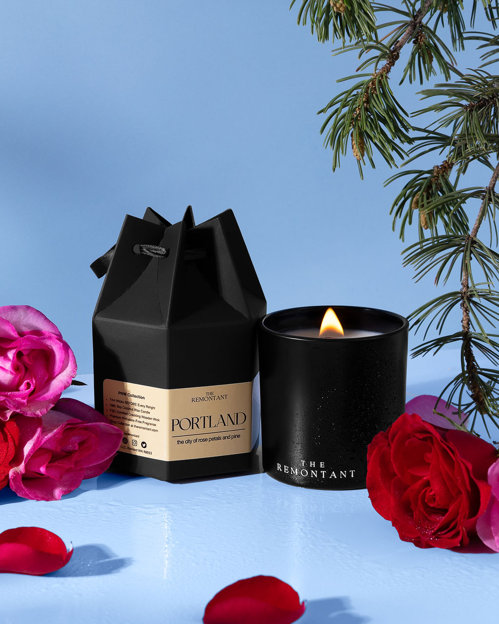 A black candle jar with a lit flame sits beside its black gift box on a light blue surface, surrounded by pink and red roses and evergreen branches. The box label reads Portland and The Remontant.