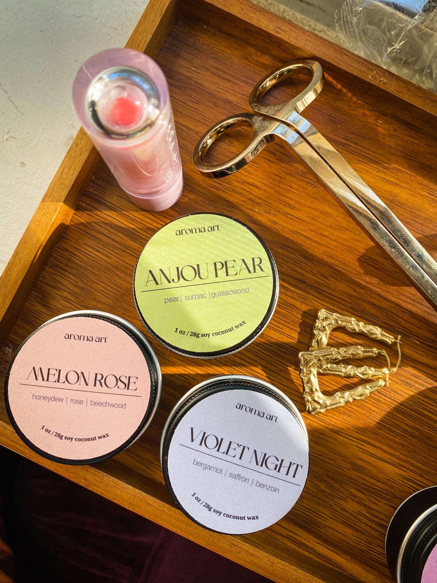 Artfully arranged on a wooden tray are travel sized tin candles in the scents Melon Rose (notes of honeydew, rose, and beechwood), Anjou Pear (notes of pear, sumac, and guaiac wood), and Violet Night (notes of bergamot, saffron, and benzoin).
