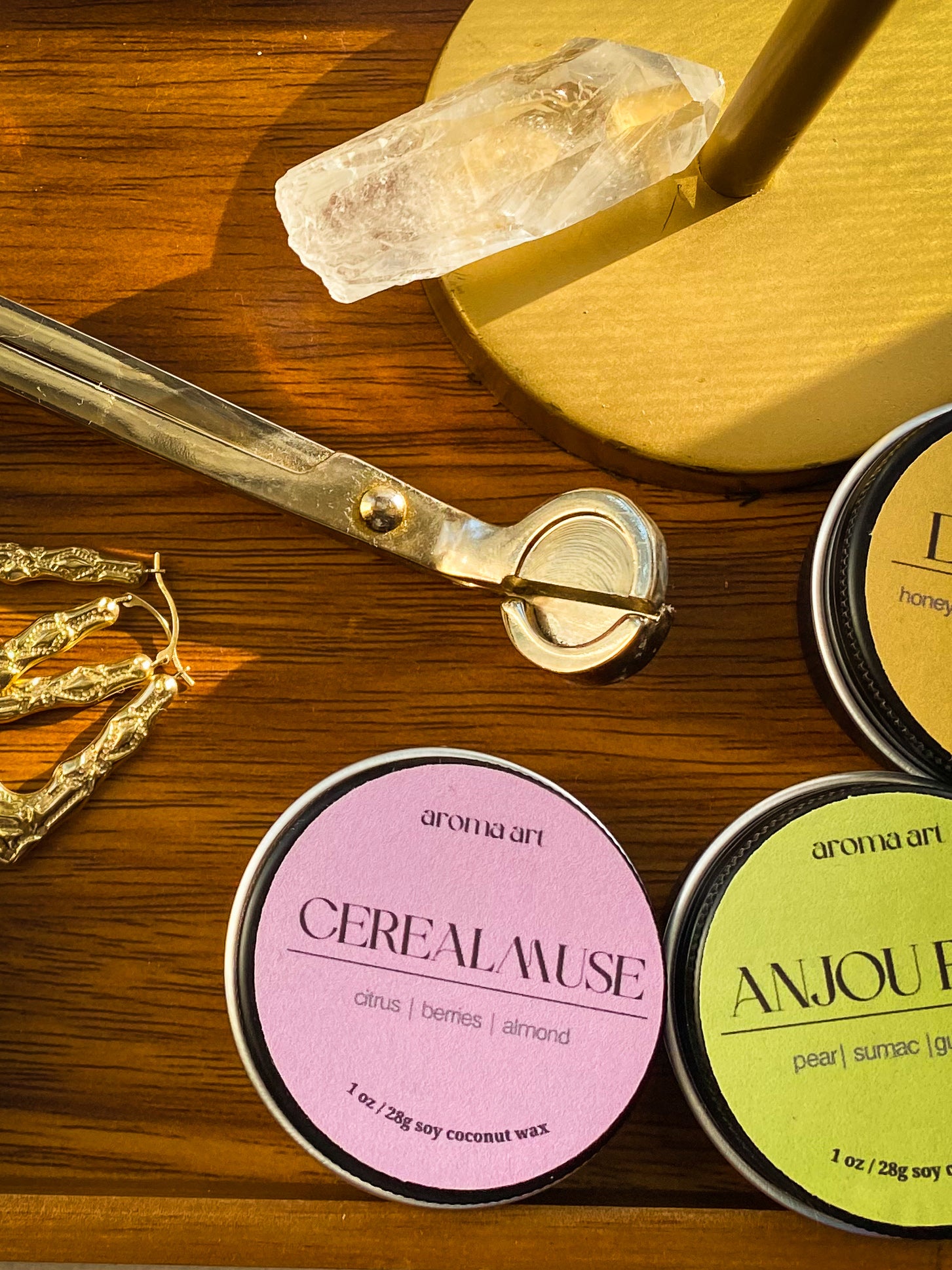 Artfully arranged on a wooden table with a wick trimmer and a crystal are travel sized tin candles in the scents Cereal Muse (notes of citrus, berries, and almond), Anjou Pear (notes of pear, sumac, and guaiac wood), and Dazzling (notes of honey, orange marmalade, and tobacco).
