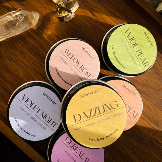 A stack of six candles in travel size tins in a variety of scents on a wooden table. The scents are (from left to right) Violet Night, Cereal Muse, Melon Rose, Dazzling, Delia Petal, and Anjou Pear. Each travel sized tin candle features a crackling wooden wick.