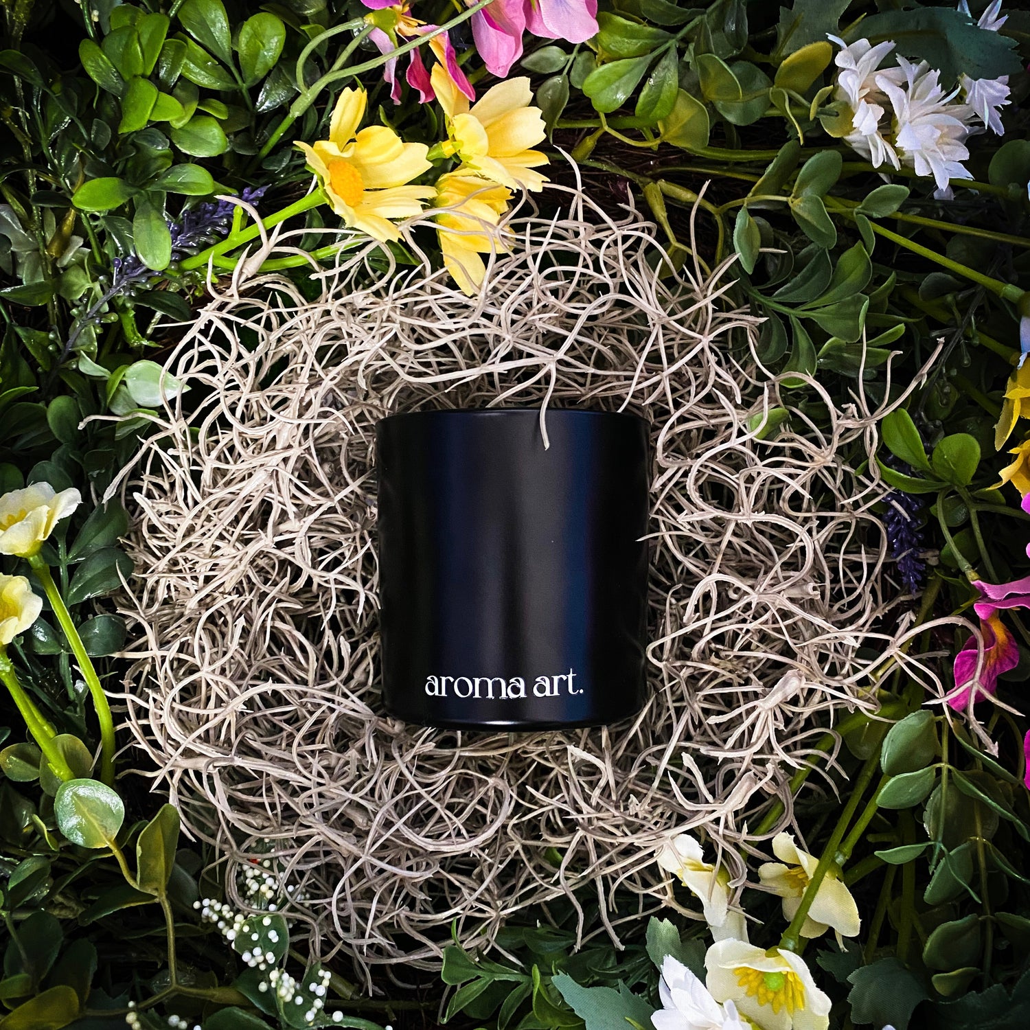 7 oz Meadow crackling wooden wick candle with notes of grass, basil, and black tea. Shown in its elegant black vessel, surrounded by nest of wildflowers. An earthy, verdant, and floral candle perfect for spring.